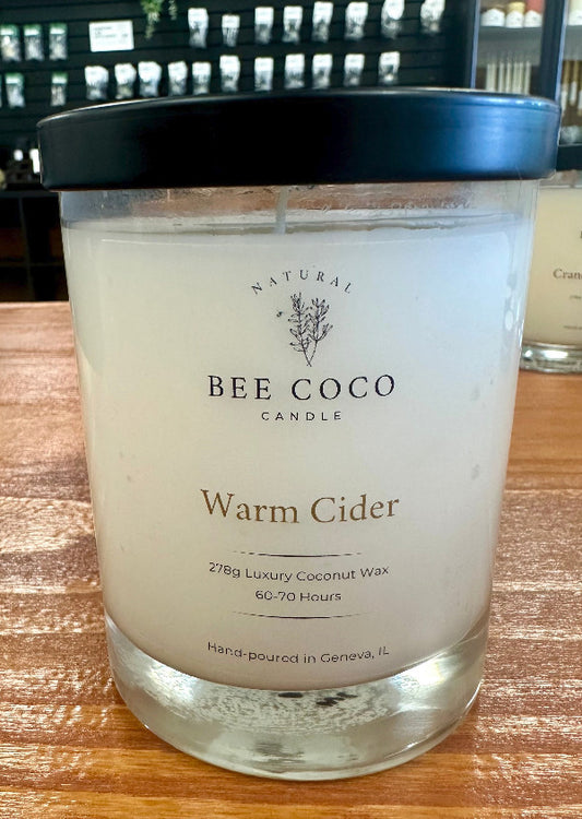 A clear glass jar candle with a label reading 'BEE COCO WARM CIDER', indicating a zesty luxury coconut wax blend with natural fragrance oils.