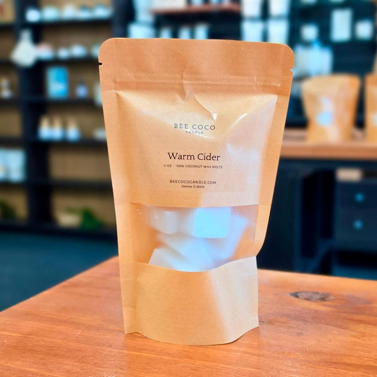 A stand-up kraft paper pouch of Bee Coco Candle Warm Cider wax melts sits on a wooden table with a softly blurred shop interior in the background.