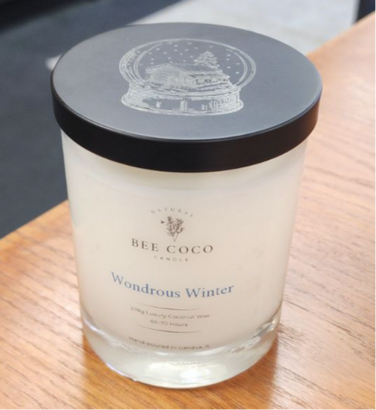 A clear candle with a black lid labeled 'Bee Coco Candle' with the scent name 'Wondrous Winter'. The candle is placed on a wooden surface.