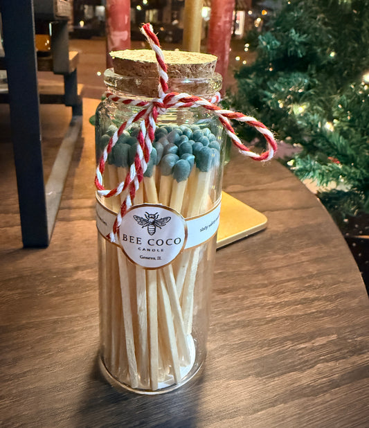 Jar of green matches with a decorative label on a wooden surface, Christmas tree in the background