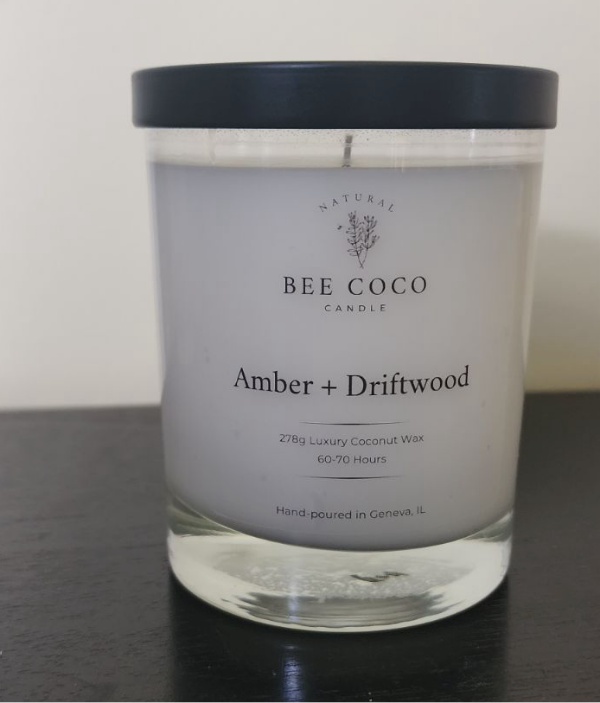A white candle in a clear glass jar with a black lid, labeled 'Amber + Driftwood', from Bee Coco Candle.