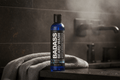 A bottle of Badass Beard Wash stands on a dark countertop with a gray towel draped beside it in a bathroom setting. The product bottle is blue with a black cap and a black label featuring white text.