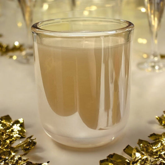 A transparent glass candle jar with a beige candle inside, placed on a table with golden decorative elements around it, likely set for a New Year's Eve celebration.