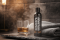 A bottle of Chuck's Hog Wash animal repellent spray stands on a dark stone surface next to a glass of amber liquor and a folded gray towel, with steam rising in a moody indoor setting.