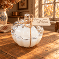 A clear glass pumpkin-shaped candle filled with white wax sits on a rustic wooden table in a warm, sunlit room. A small twine bow and paper tag are tied around the neck, with the tag reading '2026 Autumn Fest Geneva' and 'Caramel & Vanilla Caramel.