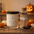 Bee Coco Candle and Match set displayed on a rustic wooden table with autumn pumpkins, leaves, and a glowing jack-o'-lantern in the background. The set includes a 'Hearth + Hollow' scented candle in a clear glass jar with a black lid and a glass jar of wooden matches with black tips wrapped in a cork and label.