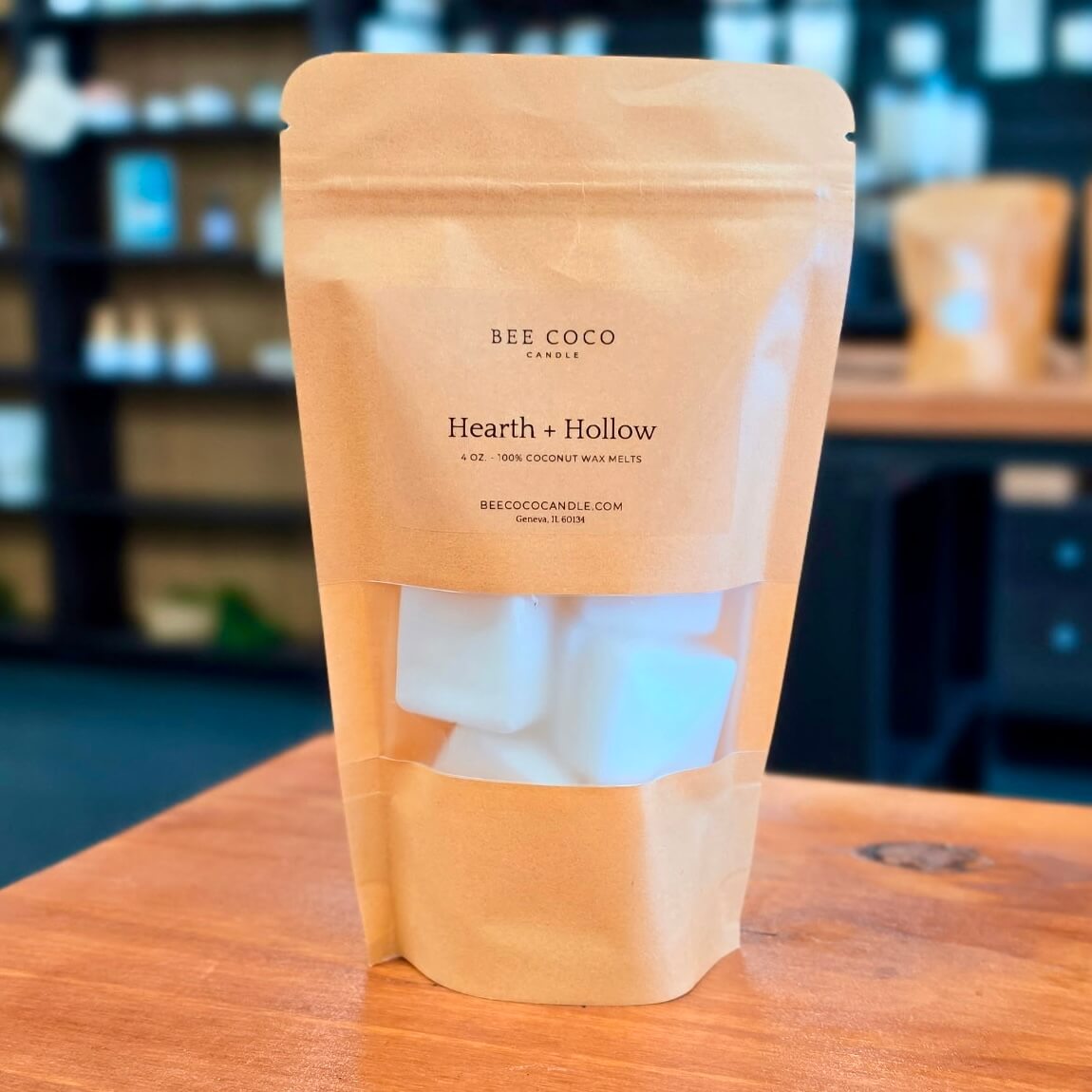A kraft paper stand-up pouch labeled 'BEE COCO CANDLES Hearth + Hollow' with a clear window showing white wax melts, standing on a wooden table in a softly blurred indoor setting.