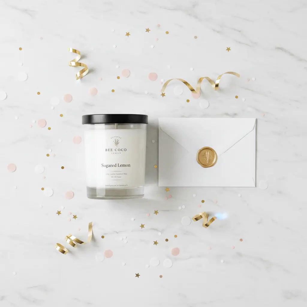 A Bee Coco Sugared Lemon candle with a black lid  beside a white envelope with a gold wax seal on a light marble surface, surrounded by gold and pink confetti and ribbon streamers.