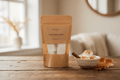 A stand-up pouch of Bee Coco Toasted Marshmallow Wax Melts sits on a rustic wooden table next to a small bowl of marshmallow toast with a cinnamon stick, with a softly blurred cozy interior background.